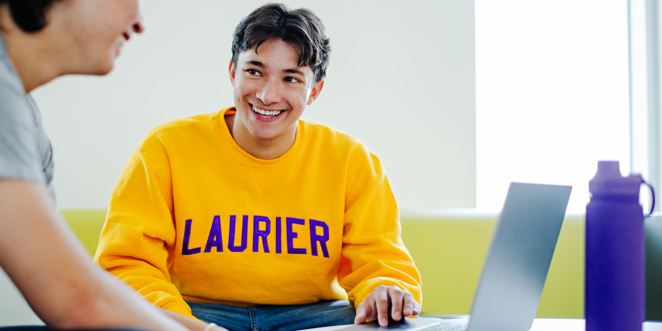 male student smiling wearing yellow Laurier hoodie on his laptop